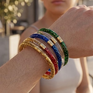 Puede incluir: Una colección de pulseras brillantes de colores oro, rojo, azul y verde, con detalles metálicos dorados. Las pulseras se llevan en una muñeca, mostrando sus colores vibrantes y su textura brillante. Un accesorio de moda.