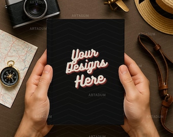 May include: A black notebook with the text "Your Designs Here" is held by hands. A vintage camera, compass, map, toy airplane, and straw hat are arranged around the notebook on a brown surface.
