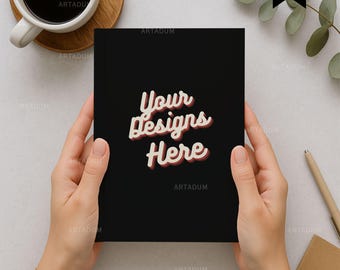 May include: A black notebook held by hands, featuring the text "Your Designs Here" in a retro font. The notebook is surrounded by a cup of coffee, a wooden coaster, a pen, and a small notepad, creating a workspace setting.