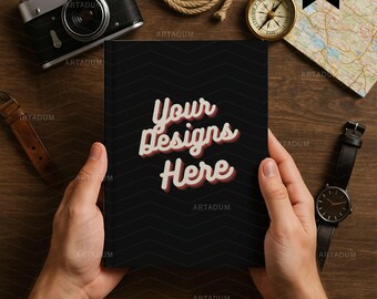 May include: A black notebook with the text "Your Designs Here" in a retro font is held by hands. A vintage camera, compass, map, rope, and watch are arranged around the notebook on a wooden surface.
