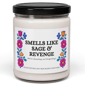 Op de afbeelding: Een witte kaars in een glazen pot met een zwart deksel. Het etiket luidt "SMELLS LIKE SAGE & REVENGE" met bloemmotieven. De tekst hieronder luidt "(We're cleansing, not forgetting.)" en "VEGAN NATURAL SOY WAX BLEND. NON-TOXIC."