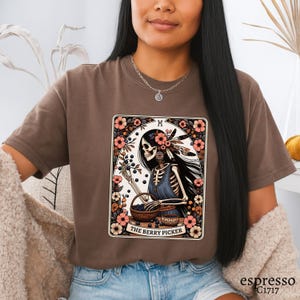 May include: Espresso-coloured t-shirt with a tarot card design. The card shows a skeleton figure with long black hair, decorated with flowers and feathers, picking berries. The text "THE BERRY PICKER" is below the image.
