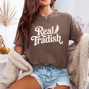 May include: A brown t-shirt with the text "Real Tradish" in a white, stylized font. A feather graphic is next to the word "Real". The shirt is worn with denim shorts and a cream-colored cardigan.