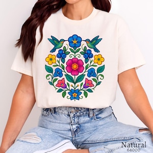 May include: Cream-colored t-shirt featuring a vibrant floral design with hummingbirds. The artwork includes a central pink flower, blue, yellow, and red blossoms, and green leaves. The text "Natural 64000" is at the bottom.