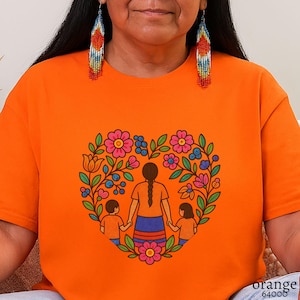 May include: Orange t-shirt featuring a heart-shaped floral design with a mother and two children holding hands. The design includes pink and blue flowers, green leaves, and a brown-haired mother figure. The text "orange 64000" is printed at the bottom right.