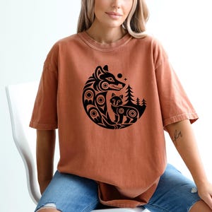 May include: A terracotta-coloured t-shirt with a black wolf and cub graphic inside a circle. The design includes trees and stars. The shirt is worn by a person sitting in a white chair.