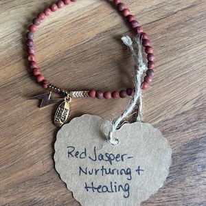 May include: Red jasper beaded bracelet with gold-colored charms, including a lightning bolt and a small tag. A heart-shaped tag reads "Red Jasper - Nurturing + Healing". The bracelet is on a wooden surface.