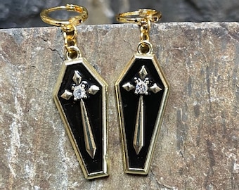 May include: A pair of gold-tone earrings with black coffin-shaped pendants. Each pendant features a gold cross with a clear rhinestone in the center.