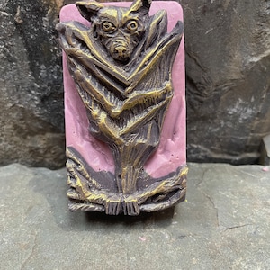 Puede incluir: Una pastilla de jabón rosa y dorada con un diseño de murciélago detallado. El murciélago está en posición de vuelo con las alas extendidas. El jabón está hecho con ingredientes naturales y es perfecto para un regalo de Halloween espeluznante.