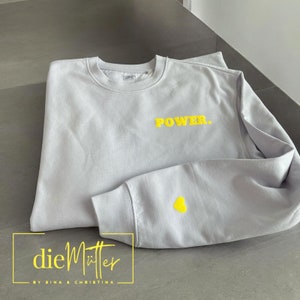 Könnte beinhalten: Ein hellgrauer Pullover mit dem Schriftzug "POWER." in gelber Schrift auf der Vorderseite und einem gelben Herz auf dem linken Ärmel. Der Pullover ist in der Hälfte gefaltet und der Markenname "dieMütter - BY SINA & CHRISTINA" ist unten sichtbar.