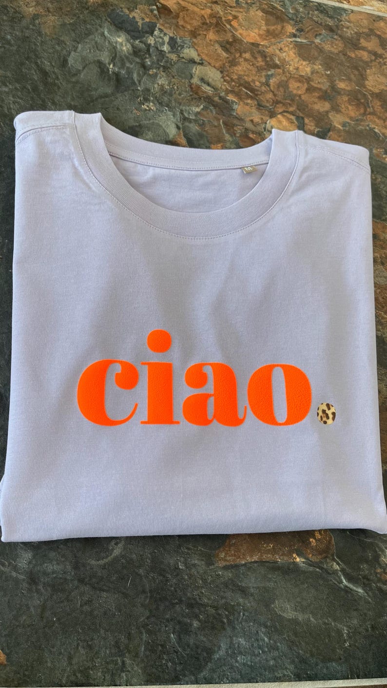 Op de afbeelding: Een lavendelkleurig T-shirt met het woord "ciao" in oranje gedrukt.