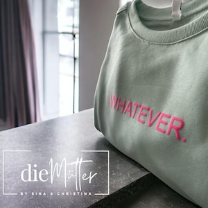 Könnte beinhalten: Ein salbeigrünes Sweatshirt mit dem Wort "WHATEVER" in leuchtendem Pink gestickt. Das Sweatshirt ist auf einer dunkelgrauen Oberfläche gefaltet. Der Markenname "die Mütter" wird in weißer Schrift angezeigt.