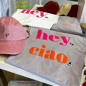Op de afbeelding: Een roze baseballpet met een klein luipaardhartje op de voorkant. De pet ligt op een tafel met verschillende grijze T-shirts met de woorden "hey ciao" erop gedrukt in roze en oranje letters. De T-shirts hebben kleine luipaardvlekken.