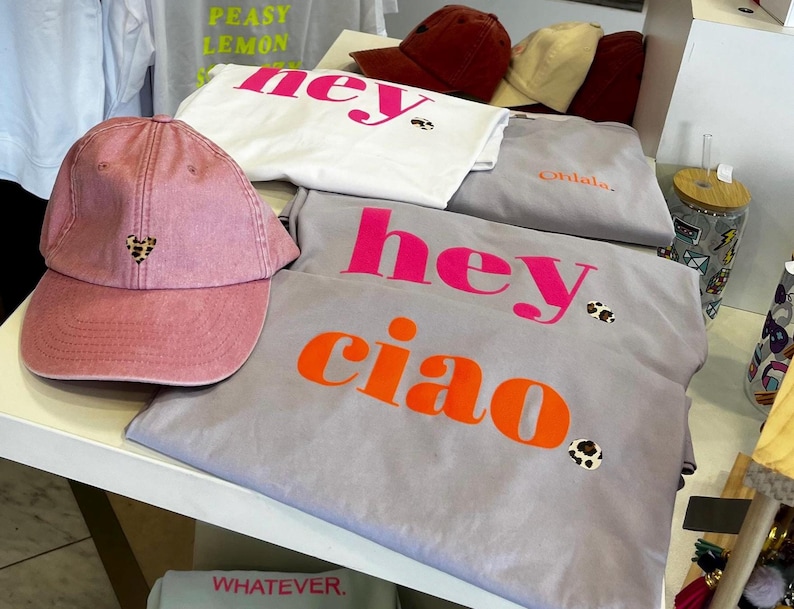 Op de afbeelding: Een roze baseballpet met een klein luipaardhartje op de voorkant. De pet ligt op een tafel met verschillende grijze T-shirts met de woorden "hey ciao" erop gedrukt in roze en oranje letters. De T-shirts hebben kleine luipaardvlekken.