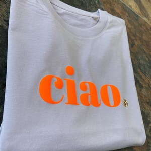 Op de afbeelding: Een lichtpaarse t-shirt met het woord "ciao" bedrukt in fel oranje.
