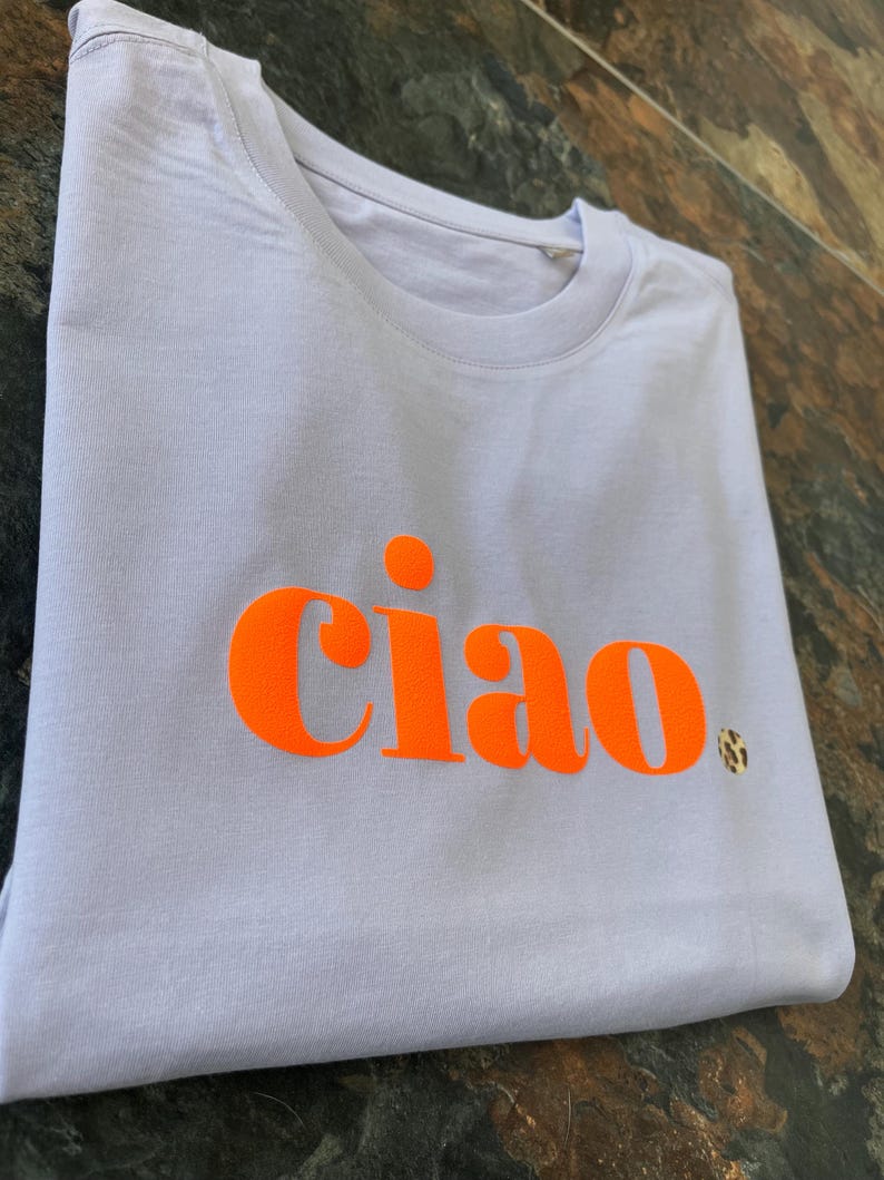 Op de afbeelding: Een lichtpaarse t-shirt met het woord "ciao" bedrukt in fel oranje.