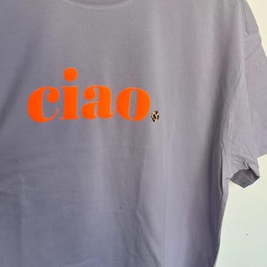Op de afbeelding: Een lavendelkleurig T-shirt met het woord "ciao" in oranje gedrukt.