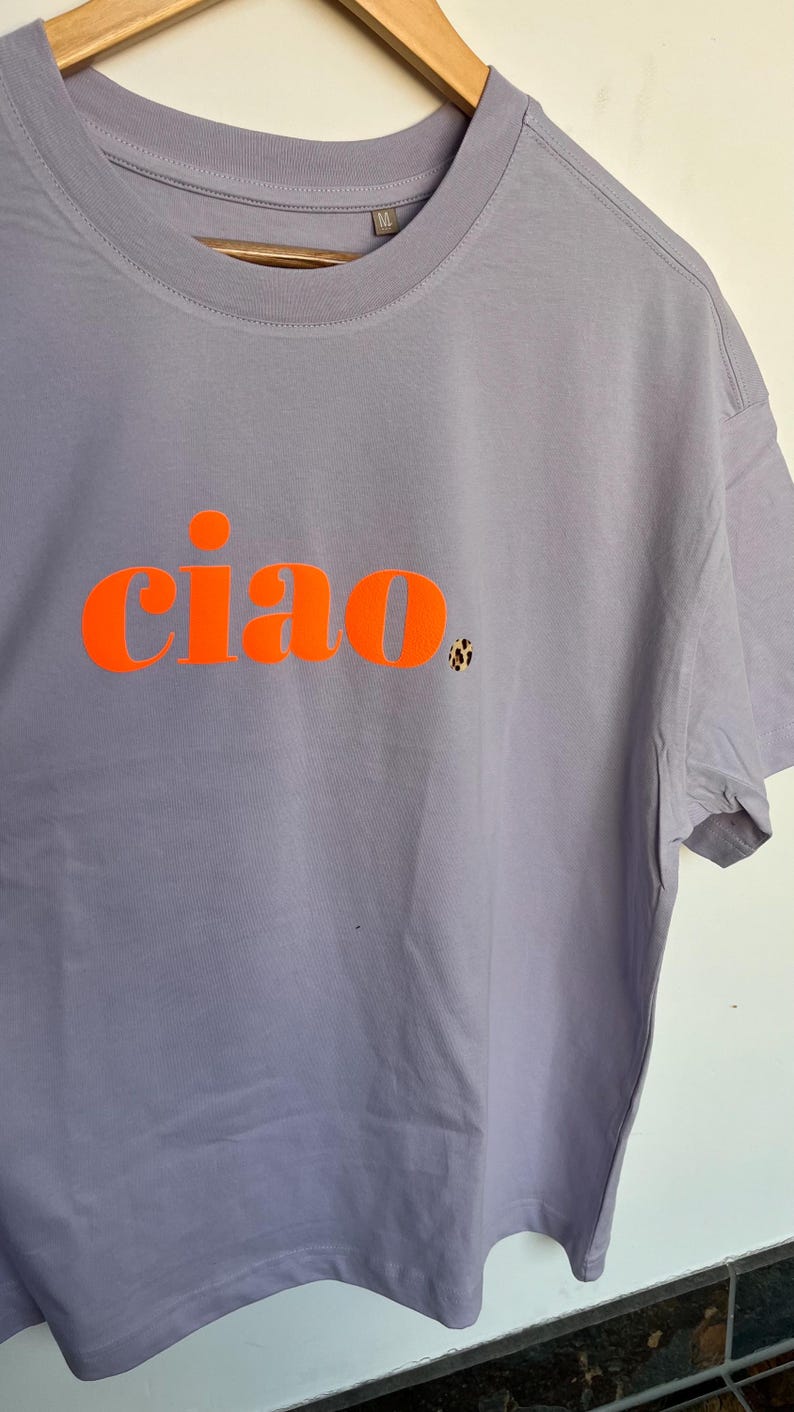 Op de afbeelding: Een lavendelkleurig T-shirt met het woord "ciao" in oranje gedrukt.