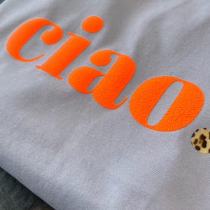 Op de afbeelding: Een lichtpaarse t-shirt met het woord "ciao" bedrukt in fel oranje. Het shirt heeft een kleine knoop met luipaardprint in de rechterbenedenhoek.