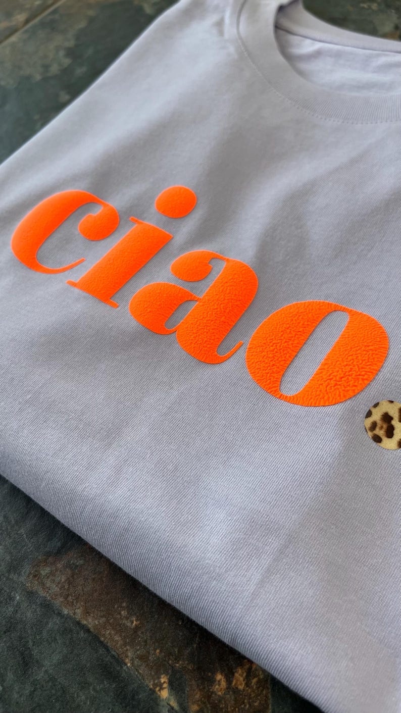 Op de afbeelding: Een lichtpaarse t-shirt met het woord "ciao" bedrukt in fel oranje. Het shirt heeft een kleine knoop met luipaardprint in de rechterbenedenhoek.