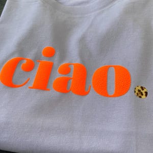 Op de afbeelding: Een lichtpaarse t-shirt met het woord "ciao" bedrukt in fel oranje. Het woord staat in een vetgedrukt, blokkerig lettertype.