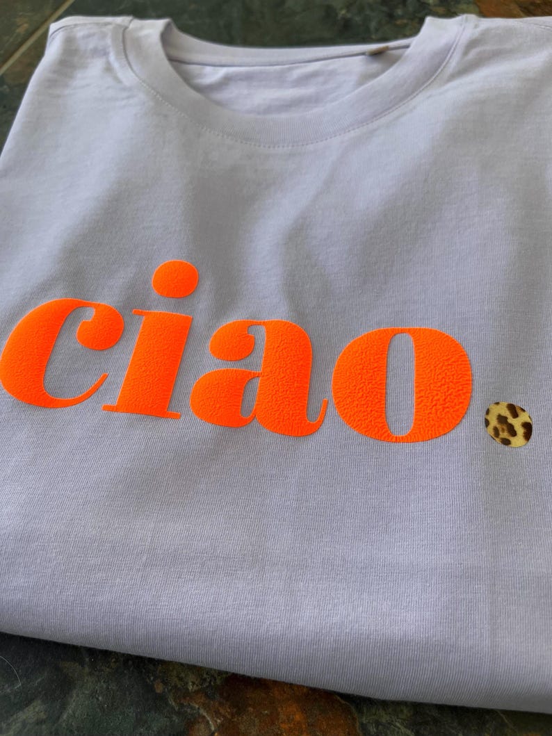 Op de afbeelding: Een lichtpaarse t-shirt met het woord "ciao" bedrukt in fel oranje. Het woord staat in een vetgedrukt, blokkerig lettertype.