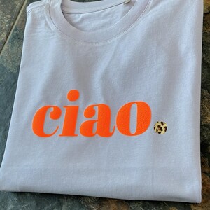 Op de afbeelding: Een lichtpaarse t-shirt met het woord "ciao" in oranje gedrukt. Een kleine sticker met luipaardprint zit aan de rechterkant van het shirt.