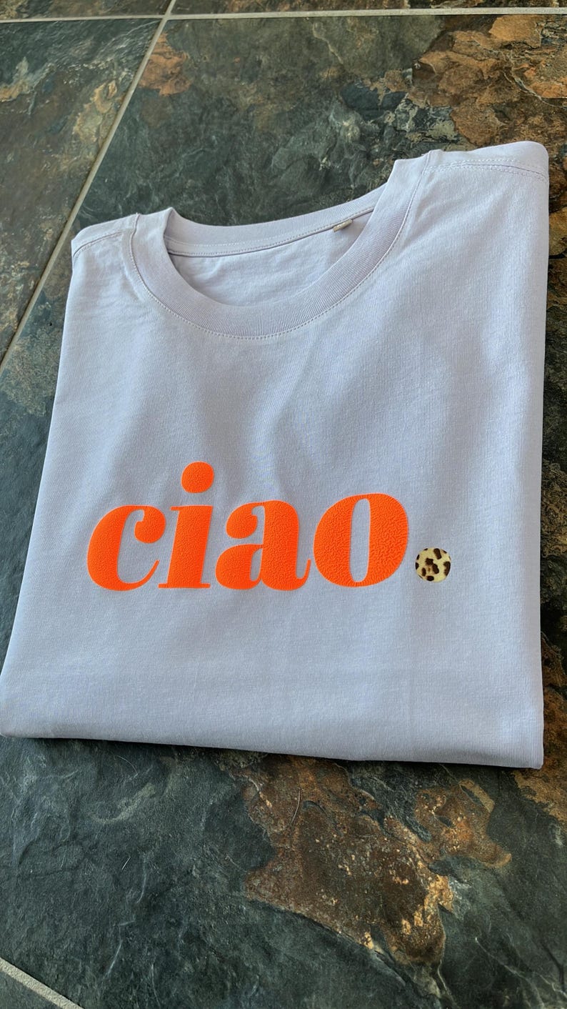 Op de afbeelding: Een lichtpaarse t-shirt met het woord "ciao" in oranje gedrukt. Een kleine sticker met luipaardprint zit aan de rechterkant van het shirt.
