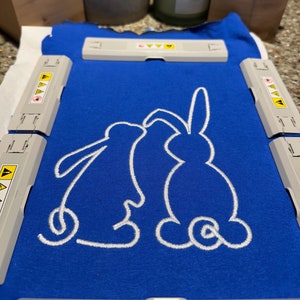 Può includere: Una maglietta blu con un disegno ricamato bianco di due conigli che stanno uno di fronte all'altro.