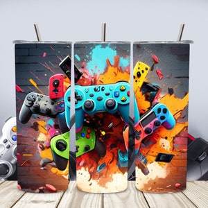 May include: A set of three colorful stainless steel tumblers with a gamer theme. The tumblers feature a variety of colorful video game controllers and joysticks against a background of orange, yellow, and blue paint splatters.