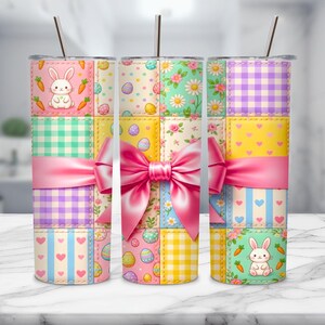 Könnte beinhalten: Drei isolierte Edelstahlbecher mit einem Ostermotiv. Jeder Becher hat ein Patchworkmuster mit Ostereiern, Hasen, Karotten und floralen Akzenten. Eine große rosa Schleife umschließt die Mitte der Becher und sorgt für einen dekorativen Touch.