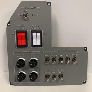Könnte beinhalten: Eine graue Steuertafel mit verschiedenen Schaltern, Knöpfen und Beschriftungen. Sie verfügt über rote und weiße Kippschalter, Drehregler und Beschriftungen wie "STBY BATT", "MASTER ALT BAT" und "LIGHTS". Das Panel ist für die Luftfahrt konzipiert.
