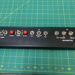 Può includere: Pannello di controllo nero con vari interruttori e pulsanti. Il pannello include interruttori a levetta contrassegnati con "LOW", "R", "L", "AUX PUMP", "START", "PRIME", "ALT", "MASTER OFF", "LEFT ENGINE", "RIGHT ENGINE" e "MAGNETOS". Sono presenti anche pulsanti rossi.