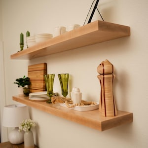 May include: Two light-colored wooden floating shelves mounted on a white wall, displaying various decorative items. Items include white dishes, green candle holders, a wooden cutting board, green glassware, and a decorative salt and pepper grinder.