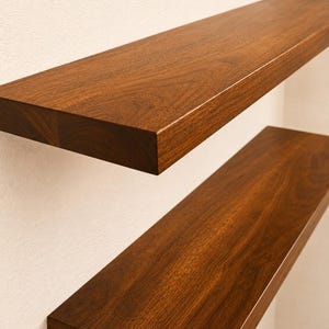 May include: Two floating shelves made of dark brown wood are mounted on a white wall. The shelves have a smooth, polished finish, and the wood grain is visible. The shelves are rectangular and appear to be sturdy.