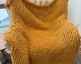 May include: A mustard yellow chunky knit blanket draped over a beige armchair. The blanket's thick, textured weave is visible, creating a cozy and inviting appearance. The blanket is made of a soft material.