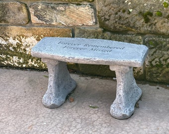 May include: A gray stone bench with the inscription "Forever Remembered Forever Missed." The bench has a rectangular seat and two tree trunk-shaped legs. It is placed on a concrete surface next to a stone wall.
