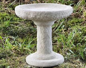 May include: A white concrete bird bath with a textured surface and a pedestal base. The bird bath is sitting on a bed of green grass.