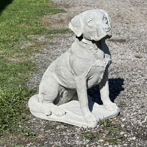 May include: A gray concrete statue of a dog sitting on a base. The dog is looking up and has a realistic appearance.