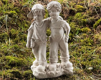May include: A light gray statue of two young boys standing close together, one with a baseball cap and a bag, the other with overalls. The statue is set on a base of stone and surrounded by grass and moss.