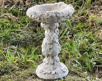 May include: A white concrete bird bath with a detailed rose design. The bath is round and sits on a pedestal with a similar rose design.