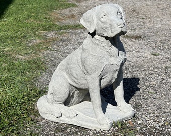 May include: A gray concrete statue of a dog sitting on a base. The dog is looking up and has a realistic appearance.