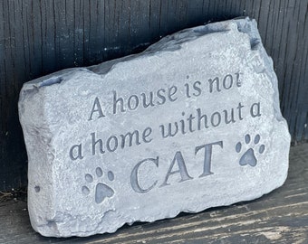 May include: A gray stone garden sign with the text "A house is not a home without a CAT" and two paw prints.