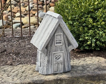 May include: A small gray concrete fairy house with a wooden door and windows. The house is sitting on a stone surface.