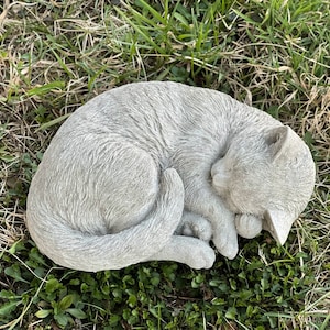 May include: A gray concrete garden statue of a cat curled up sleeping. The cat is lying on its side with its head resting on its paws. The statue is detailed with fur and facial features.