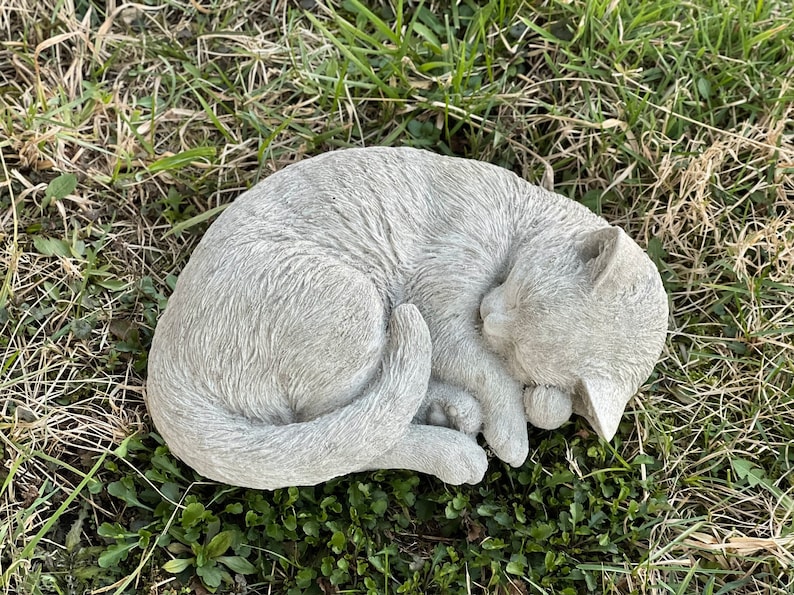 May include: A gray concrete garden statue of a cat curled up sleeping. The cat is lying on its side with its head resting on its paws. The statue is detailed with fur and facial features.