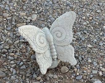 May include: A detailed, light gray concrete butterfly sculpture with intricate spiral and floral wing patterns. The butterfly is resting on a bed of small rocks and pebbles, showcasing its decorative design. A garden ornament.