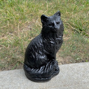Peut inclure: Une figurine de chat noir avec une queue touffue, assise sur une surface en b&eacute;ton gris.