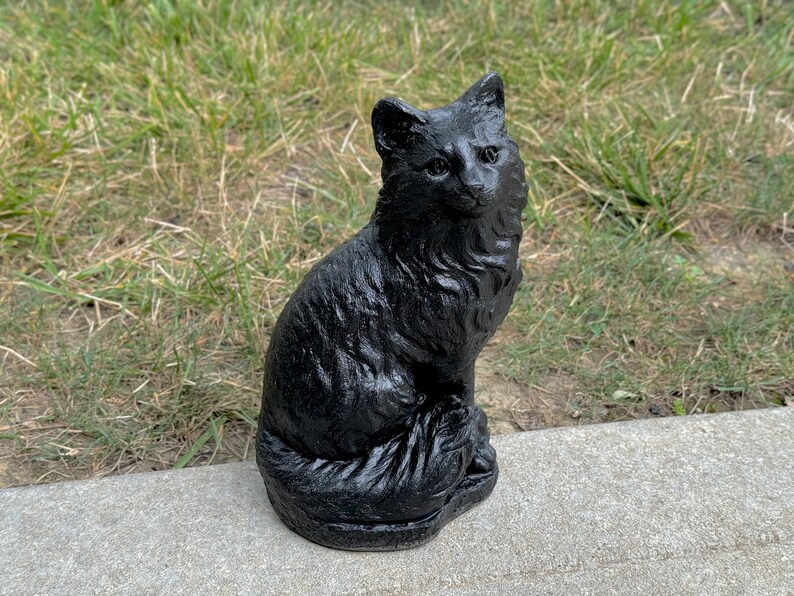 Peut inclure: Une figurine de chat noir avec une queue touffue, assise sur une surface en b&eacute;ton gris.