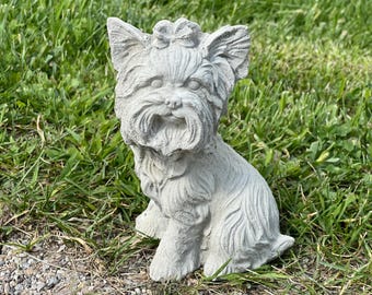 May include: A gray concrete statue of a Yorkshire Terrier dog sitting with its head tilted slightly to the side. The dog has a bow on its head and is detailed with fur and facial features.